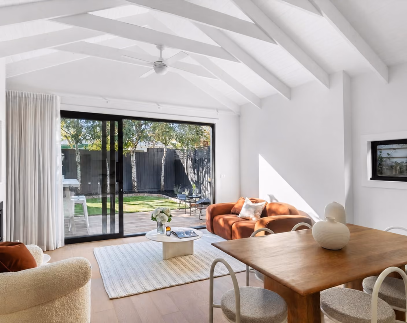 Bright living room with white ceiling beams, large sliding glass door, brown couch, and wooden dining table.