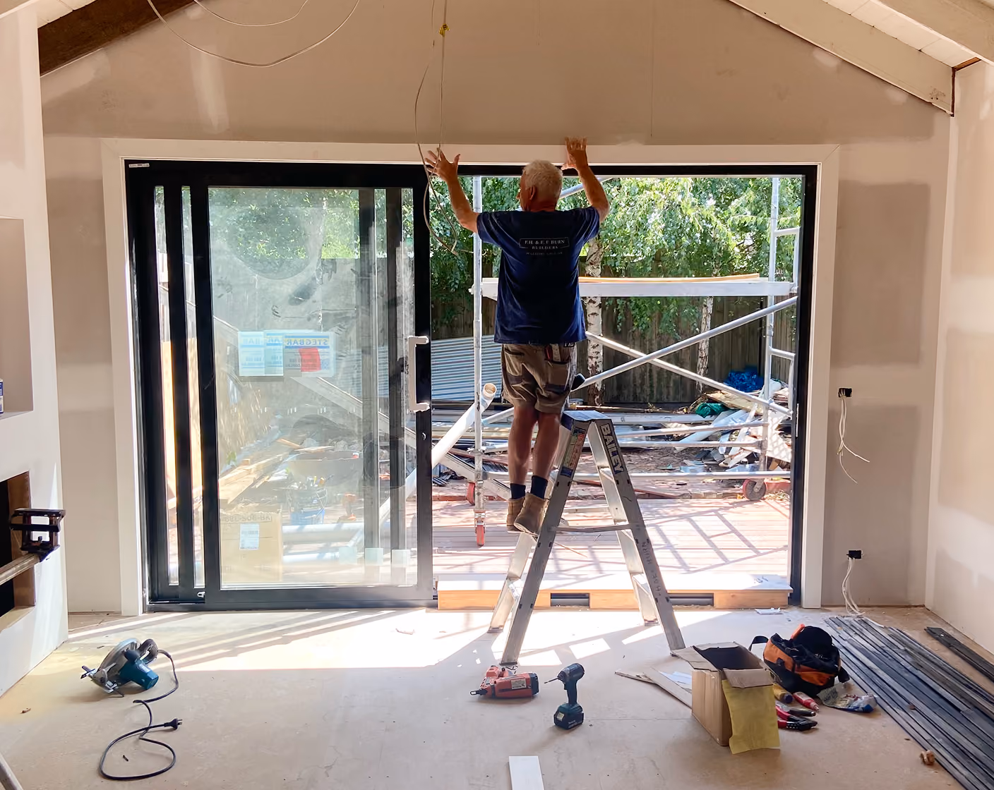 Worker on a stepladder installs or adjusts wiring near large sliding glass doors in a home under construction.
