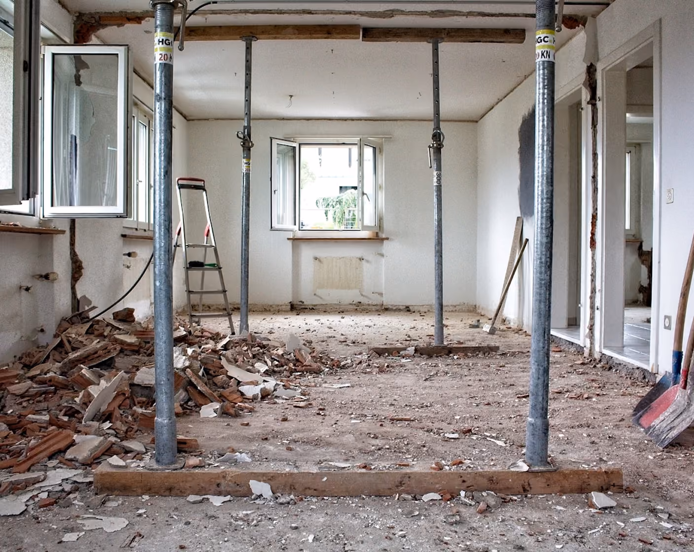 Interior room under renovation with debris, metal support poles, open windows, ladder, and construction tools.