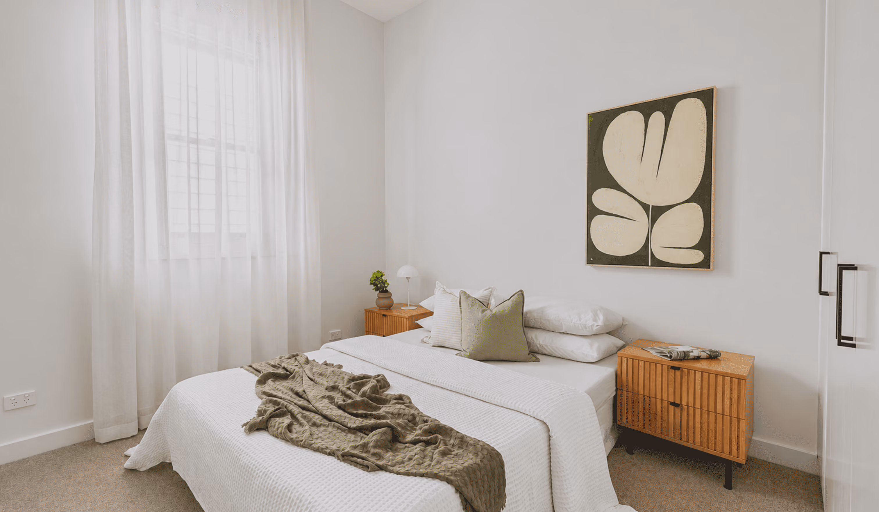 Minimalist bedroom with a bed, white bedding, a green throw, two wooden nightstands, and abstract wall art.