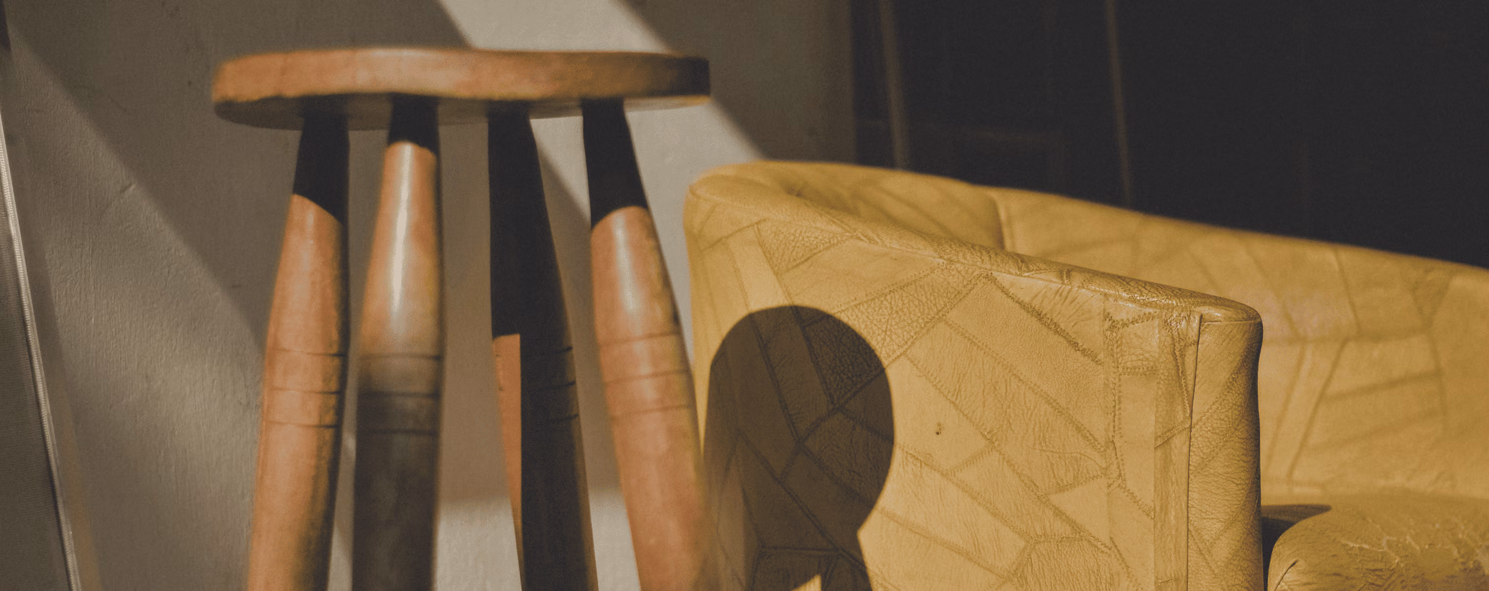 Wooden stool casting shadow on yellow leather armchair in soft sunlight indoors.