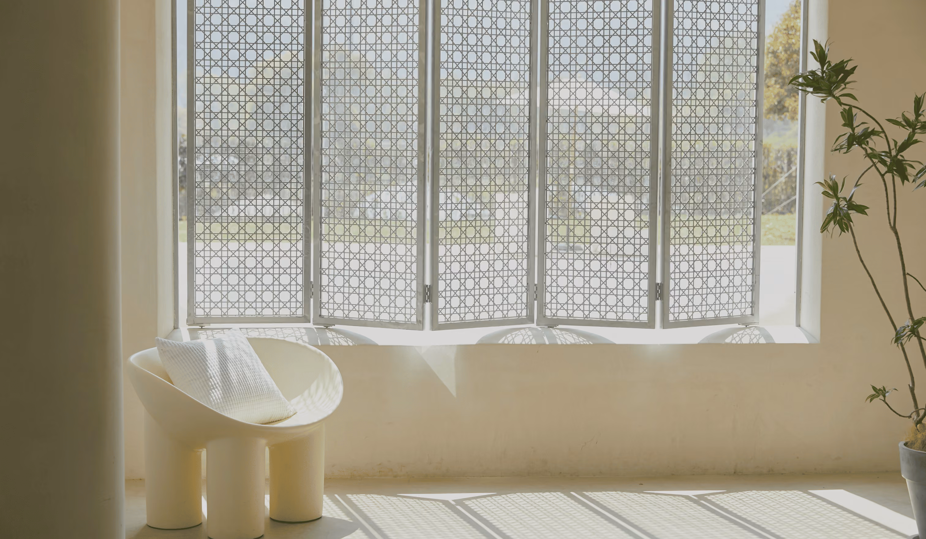 Modern white chair with cushion by window with patterned shutters casting shadows and a potted plant nearby.