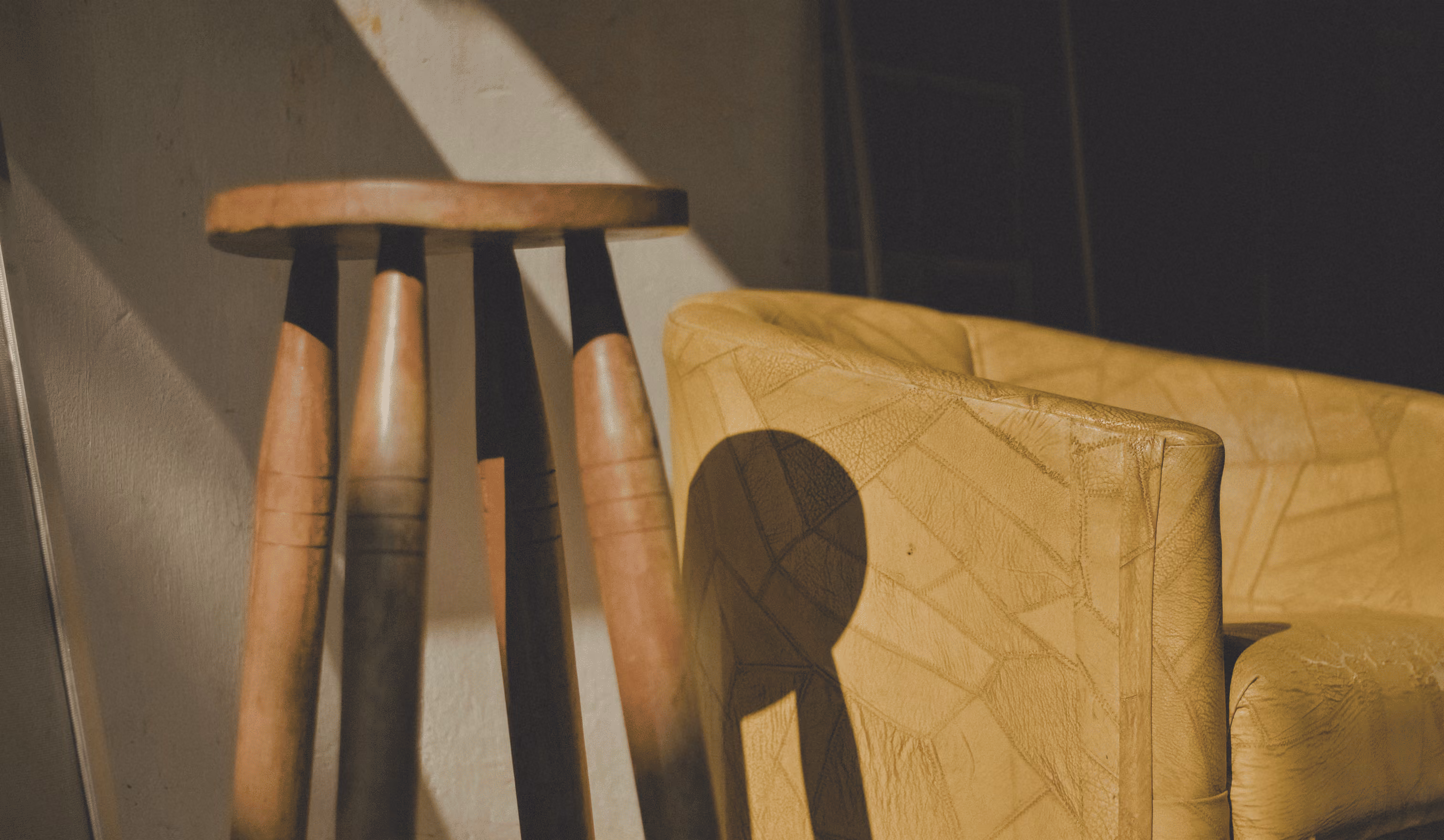Sunlight casts shadows on a wooden stool and a worn yellow leather armchair on a textured wall.