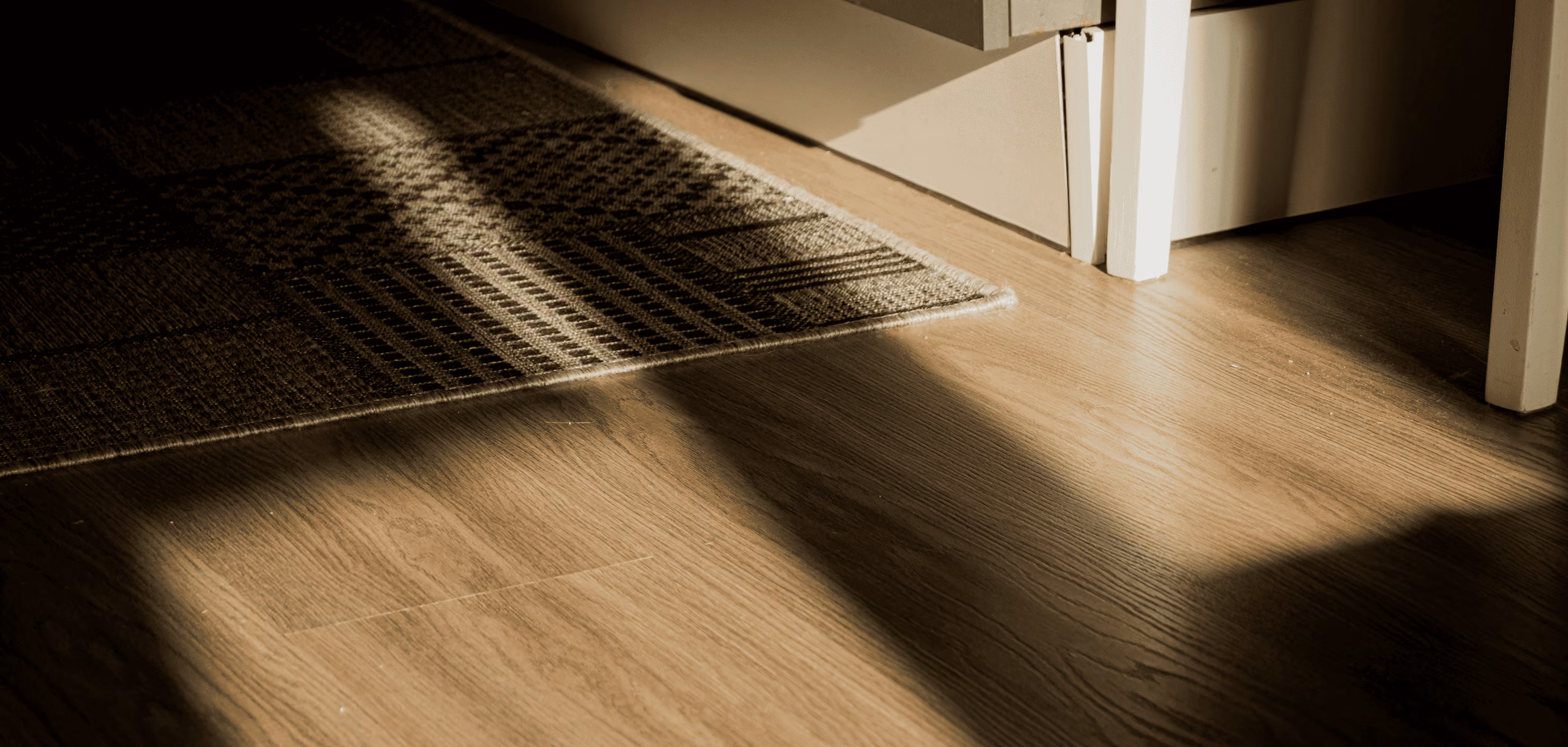 Sunlight casting shadows on a wooden floor near a patterned rug and white furniture legs.