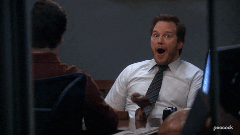 Man in white shirt and tie excitedly reacting with wide open mouth while sitting at an office desk across from another person.