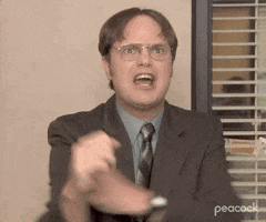 Man in glasses and a suit shaking his fist angrily while seated in an office.