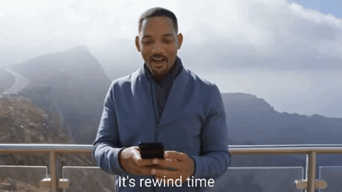 Man standing outdoors on a balcony in mountainous area, holding a phone and smiling with text 'It's rewind time' at the bottom.