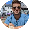 Man wearing sunglasses and a blue t-shirt sitting outdoors at a table with buildings and cars in the background.