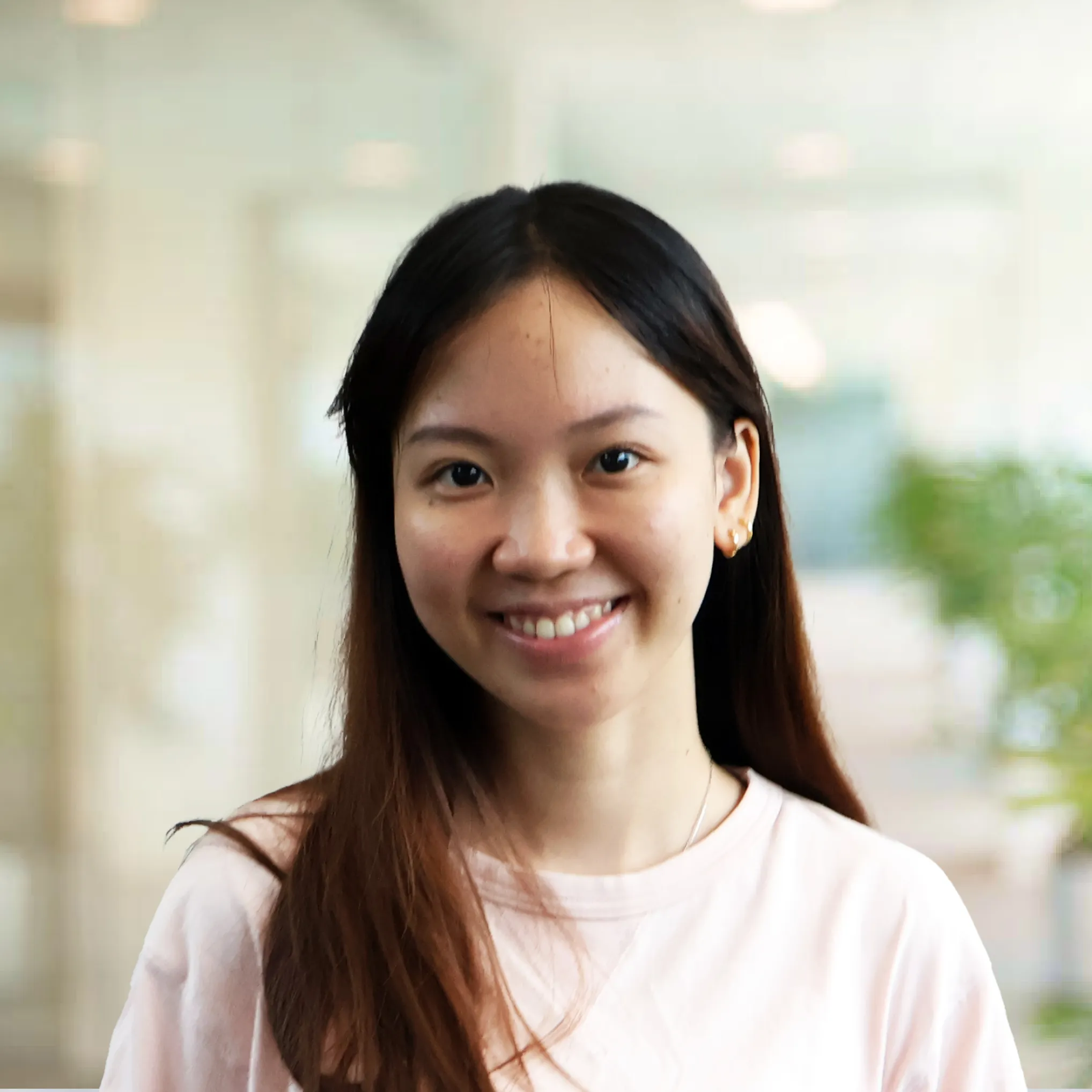 Smiling young woman with long dark hair wearing a light pink shirt in a bright indoor setting.