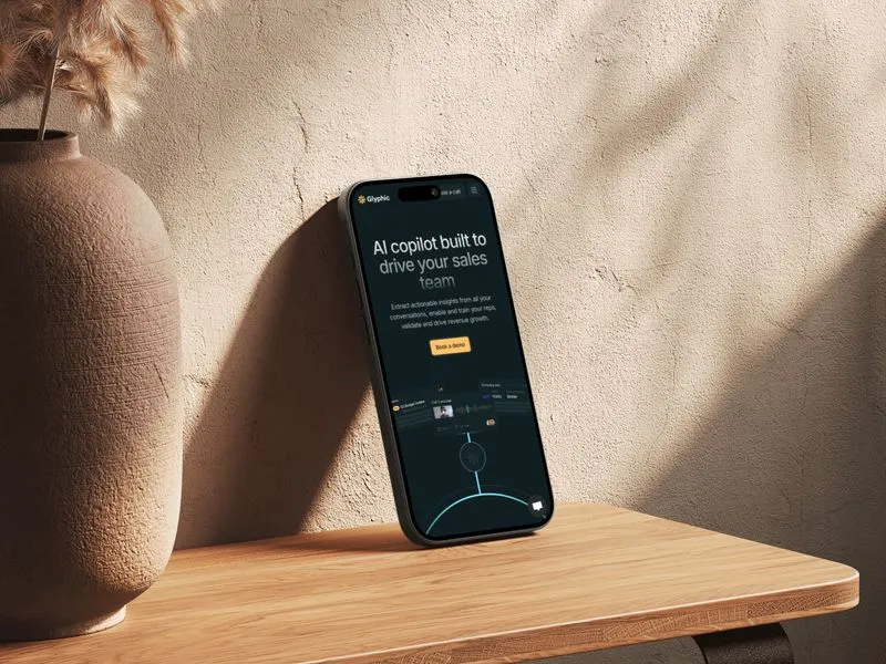 Smartphone leaning against a beige textured wall on a wooden surface, displaying a webpage titled 'AI copilot built to drive your sales team' with a 'Book a demo' button.