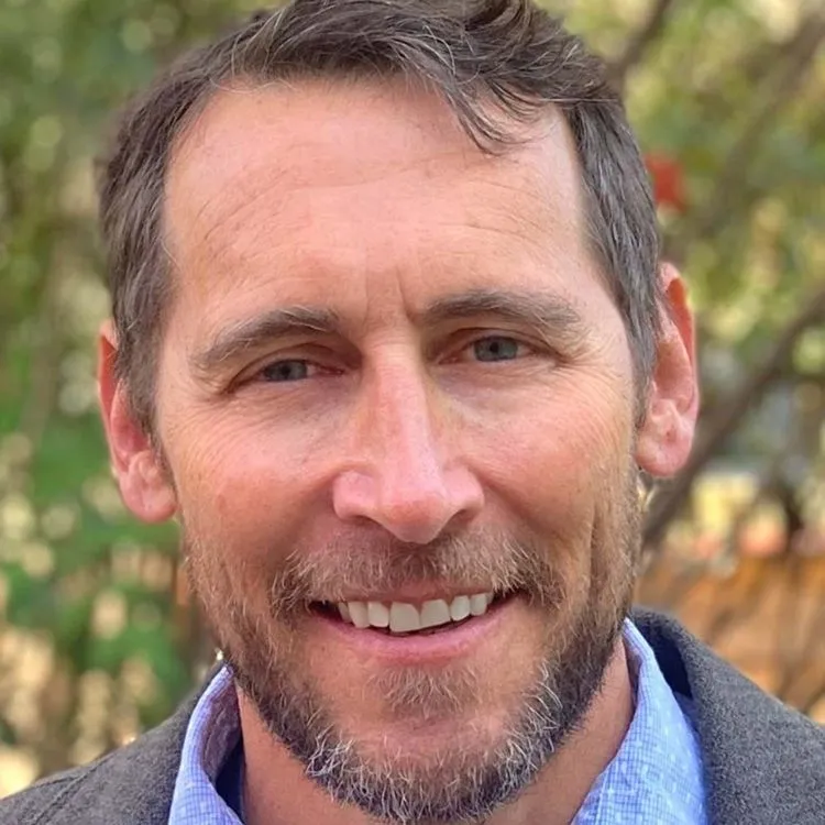 Smiling middle-aged man with blue eyes, short brown hair, and a beard wearing a blue shirt and dark jacket with a blurred outdoor background.