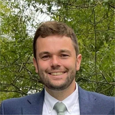 Smiling man wearing a blue suit jacket, white shirt, and light green tie standing in front of leafy green trees.