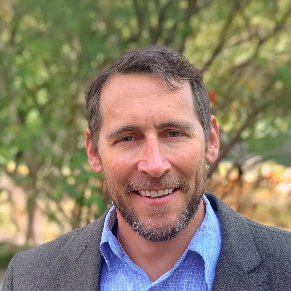 Smiling man wearing a blue suit jacket, white shirt, and light green tie standing in front of leafy green trees.