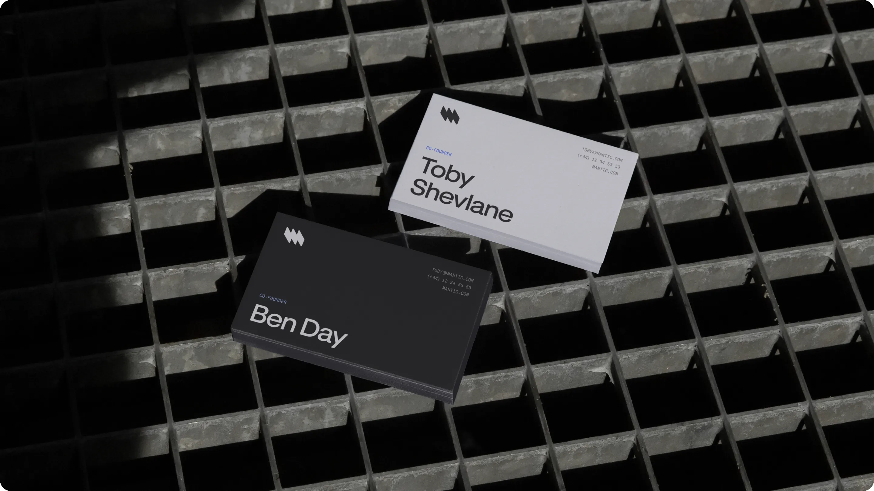 Two business cards on a metal grid surface, one black with 'Ben Day' and one white with 'Toby Shevlane', both co-founders with contact info and logo.