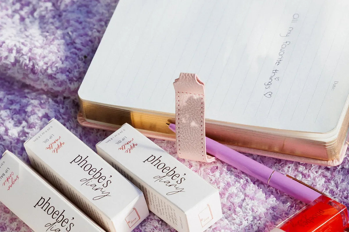 Phoebe’s Diary lip product boxes beside an open notebook and pen on a lilac knit blanket
