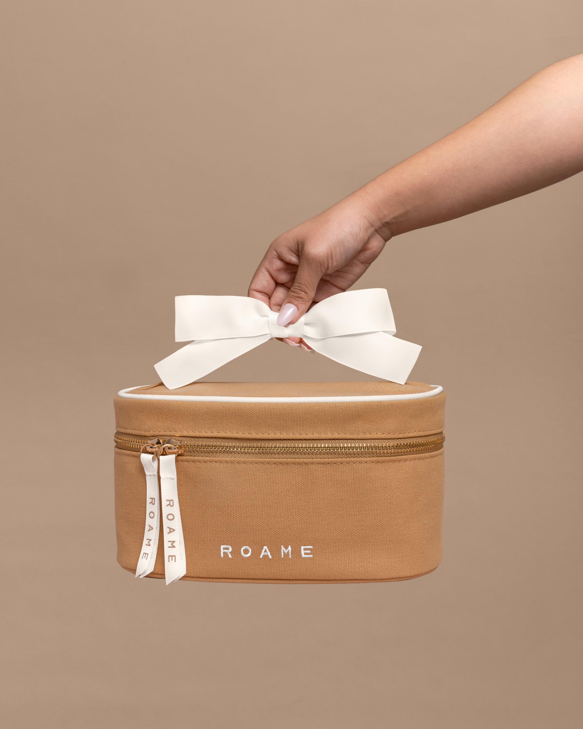 A hand lifting a tan ROAME travel case by its ribbon bow