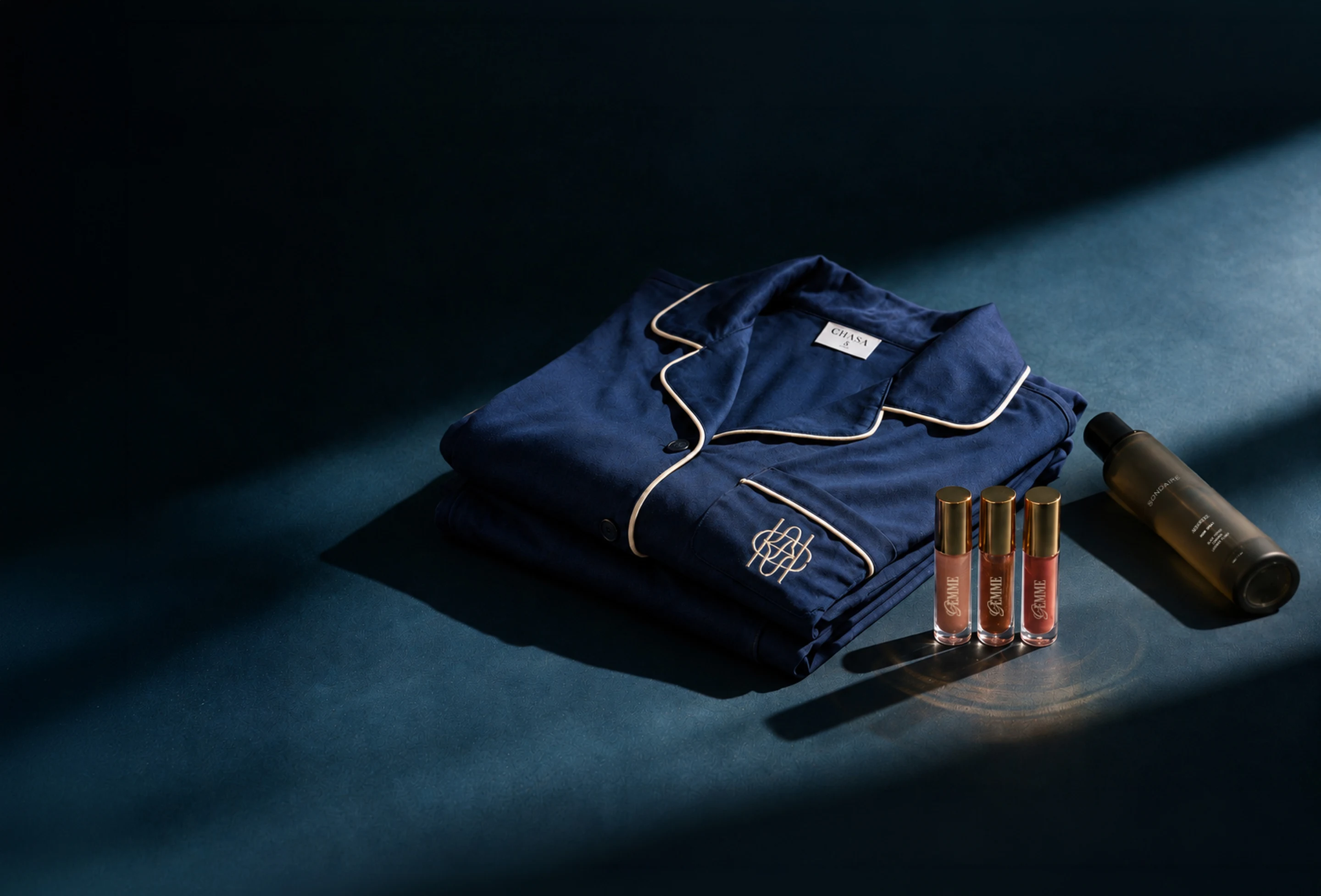 Navy monogrammed pyjamas, a room spray and three lip toppers lit on a dark surface