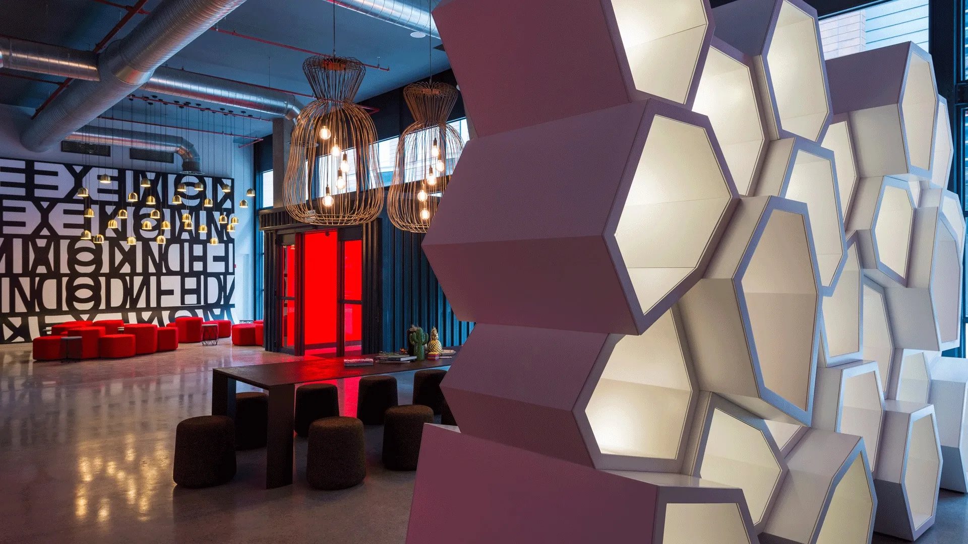 Contemporary hotel lobby with large white illuminated hexagonal wall panels, industrial-style ceiling with exposed ducts, multiple hanging cage light fixtures, long dark table surrounded by round stools, red seating cubes near a wall with artistic typographic design, and a bright red glass entrance door.