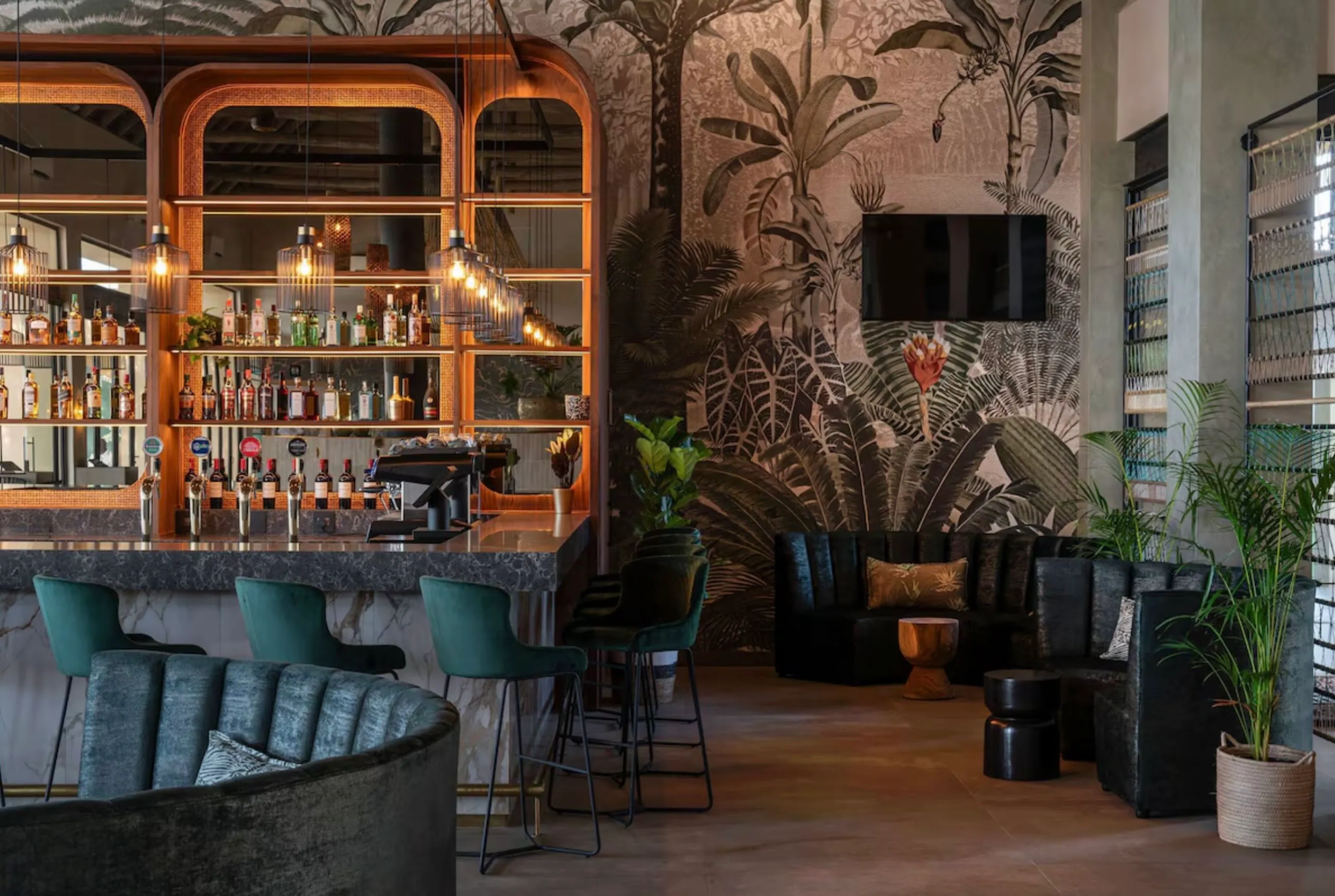 Modern bar and lounge area with green velvet bar stools at marble counter, backlit shelves with liquor bottles, pendant lights, tropical leaf mural, dark tufted seating, wooden and black stools, and potted plants.
