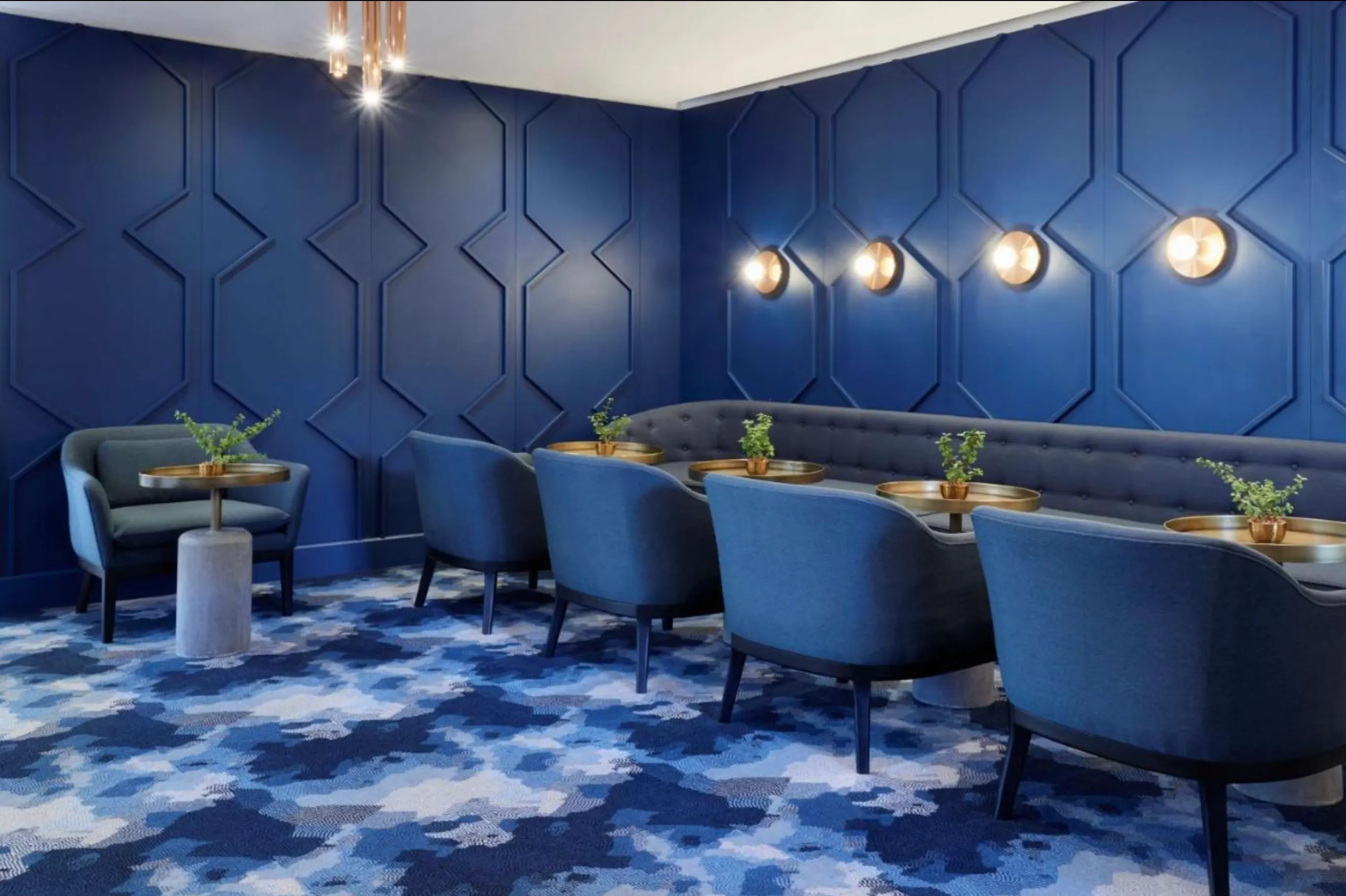 Modern lounge with deep blue geometric paneled walls, blue upholstered chairs around round wooden tables, a long tufted blue sofa, gold wall sconces, and a patterned blue carpet.