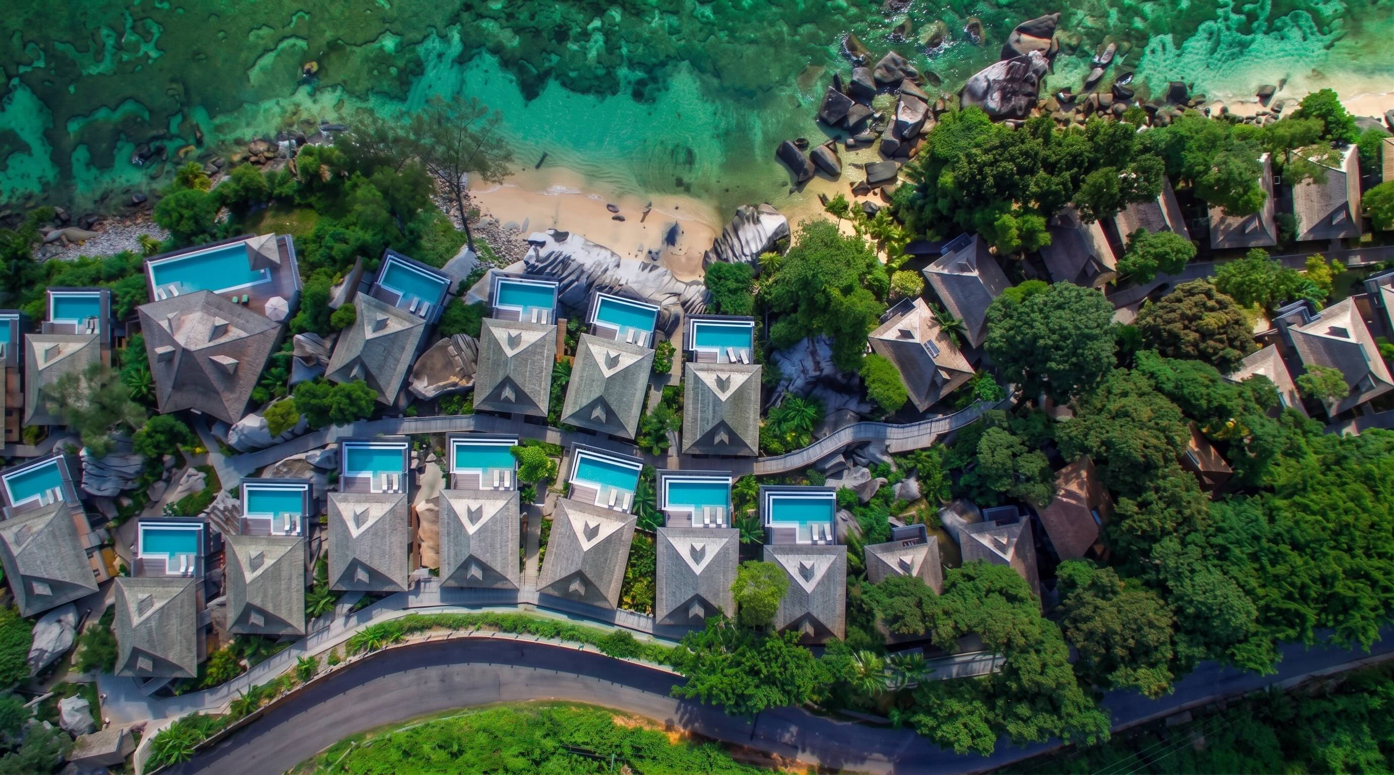 Aerial view of a tropical resort with multiple villas featuring private swimming pools, surrounded by lush green trees, a sandy beach, clear turquoise water, and a curved road at the bottom.