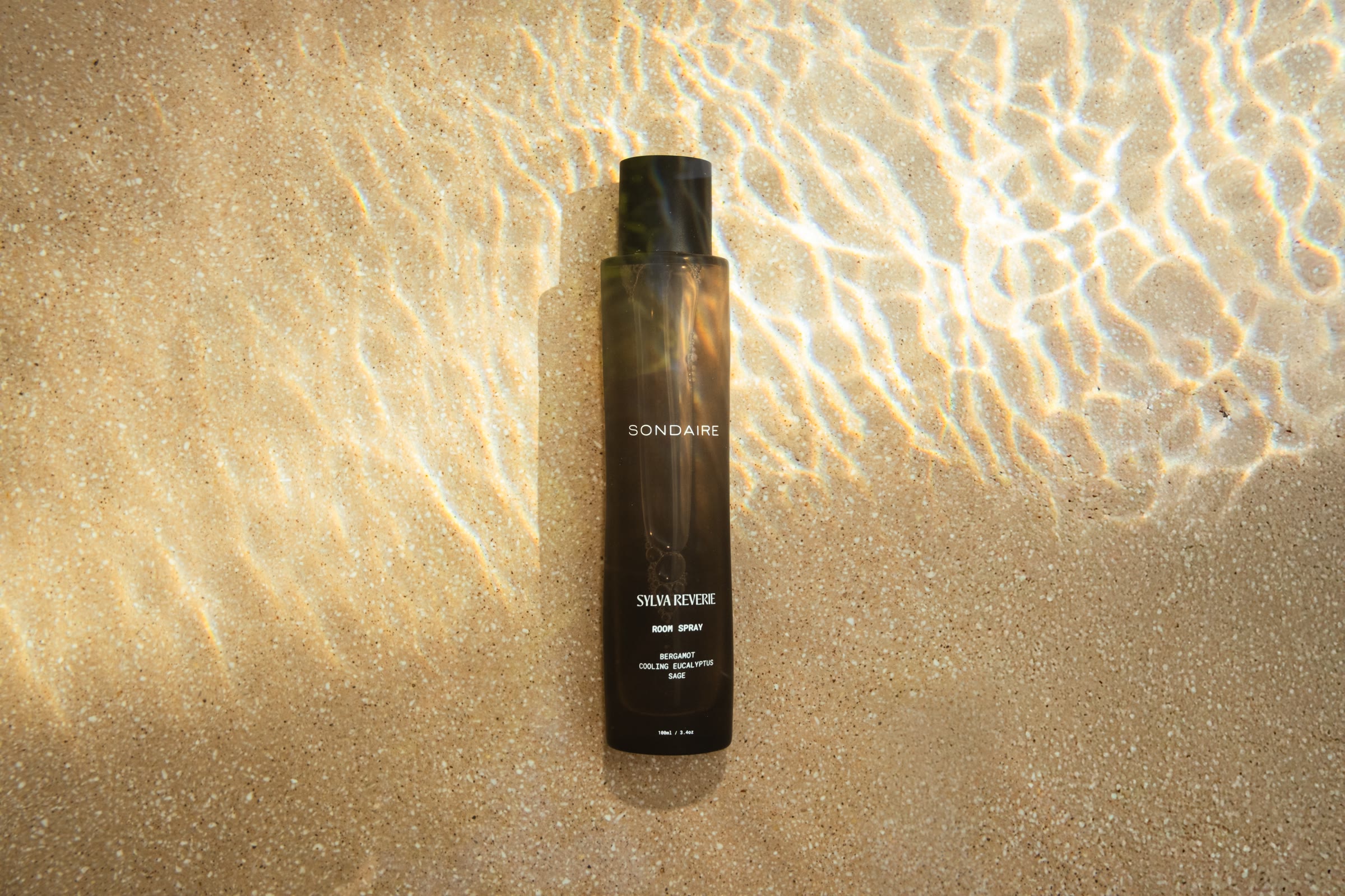 A Sondaire room spray bottle resting on wet sand at the shoreline