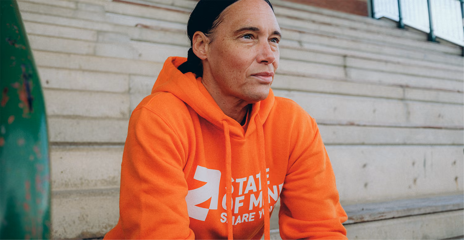 Person with dark hair and a black headband wearing an orange hoodie sitting on outdoor concrete steps, looking thoughtfully to the right.