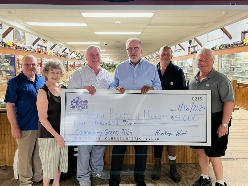 Six adults standing indoors in a hallway lined with display cases and model trains, holding a large ceremonial check for $4,000 dated 7/16/2024, payable to Medina Railroad Museum from Heritage Wind labeled Community Grant 2024.