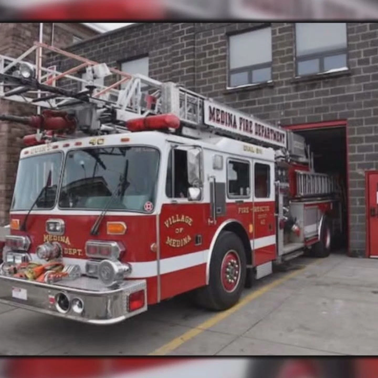 Red and white fire truck with an extended ladder parked outside a brick fire station building, labeled 'Medina Fire Department' and 'Village of Medina'.