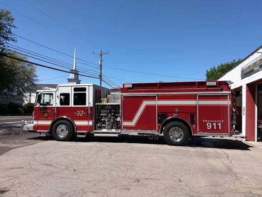 Red and white fire truck parked outside a fire station on a sunny day with a clear blue sky and a church steeple in the background.