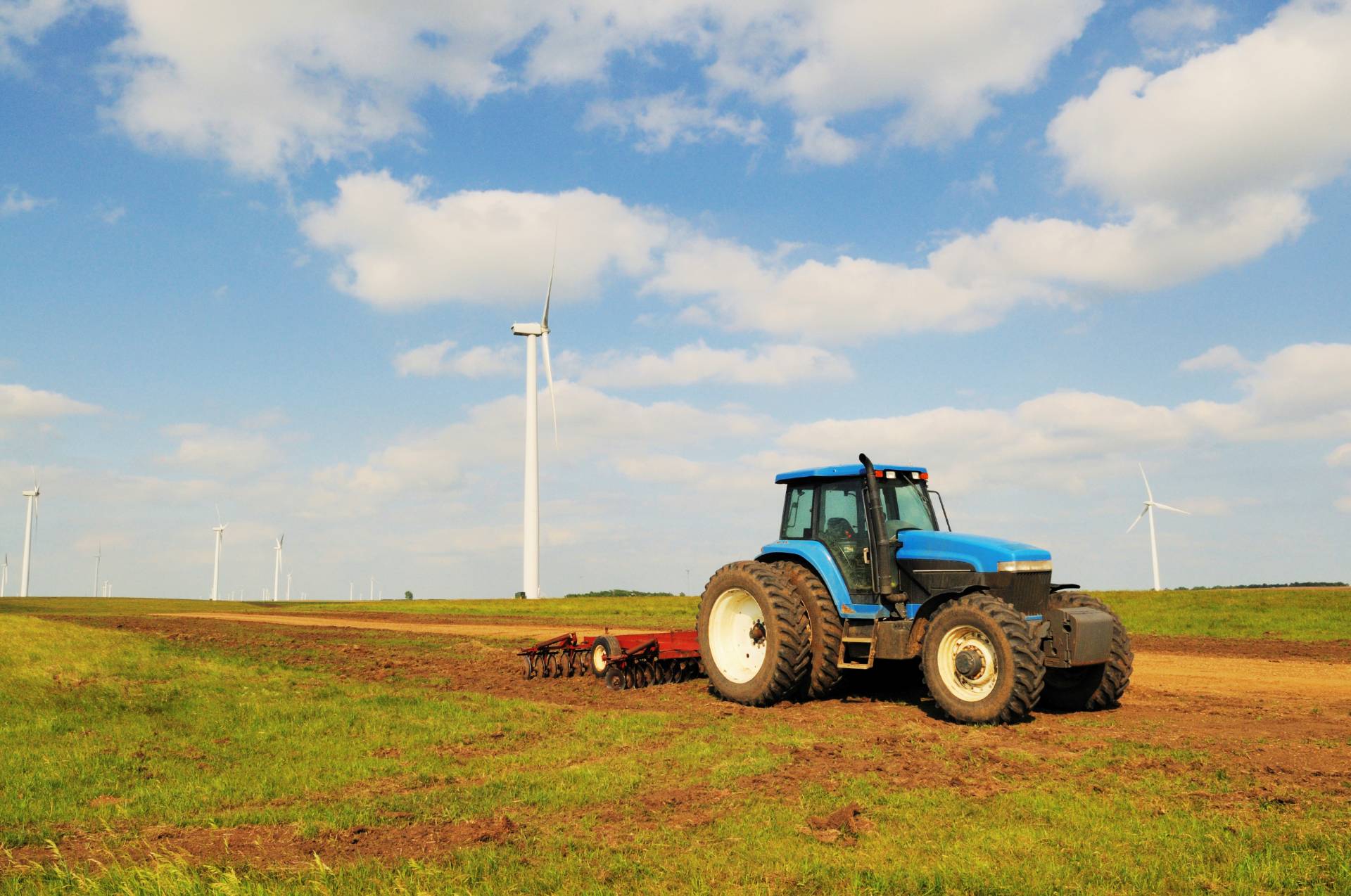 Blue tractor with farming equipment on green field with wind turbines under a partly cloudy sky.