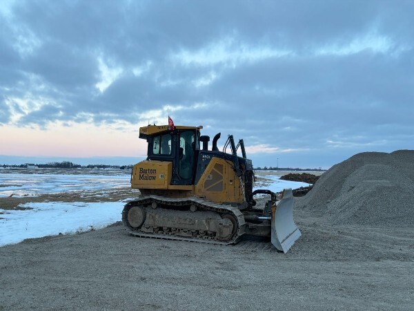 Yellow bulldozer labeled Barton Malow parked on dirt next to a large pile of earth under a cloudy sky.