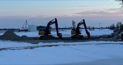 Two excavators working on snow-covered construction site with piles of dirt and large pipes nearby.
