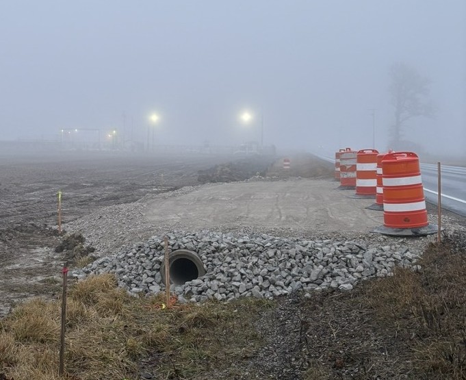 Foggy scene of a construction site beside a road with orange and white traffic barrels lined up, gravel-covered drainage pipe opening in the foreground, and dim streetlights hazily glowing in the distance.