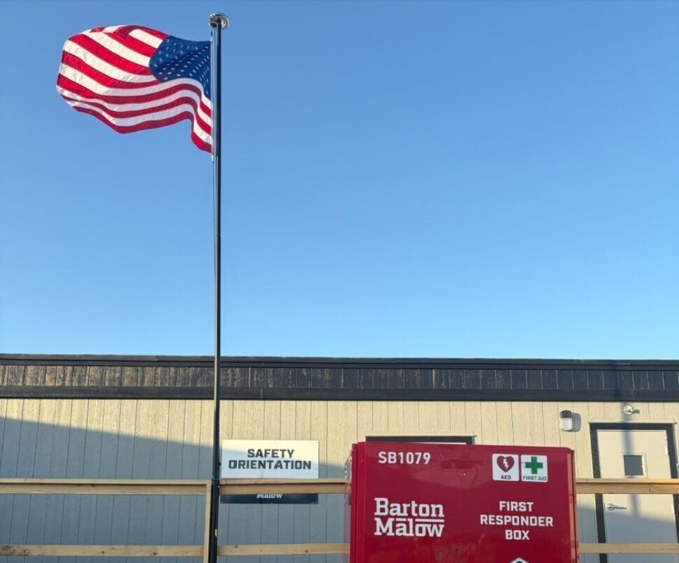 American flag flying on a tall flagpole against a clear blue sky above a beige and brown building with a safety orientation sign and a red Barton Malow first responder box.
