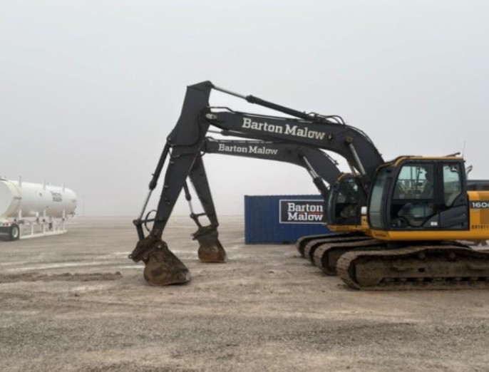 Two large tracked excavators with Barton Malow branding parked side by side on a gravel surface with a foggy background, next to a blue container also marked Barton Malow.