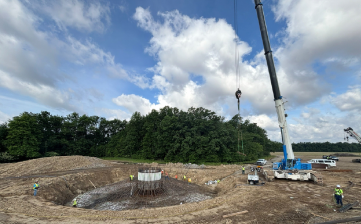 Construction site with workers assembling a large circular concrete foundation reinforced with steel rebar under a partly cloudy sky, surrounded by dirt and trees, with a tall crane lifting materials.