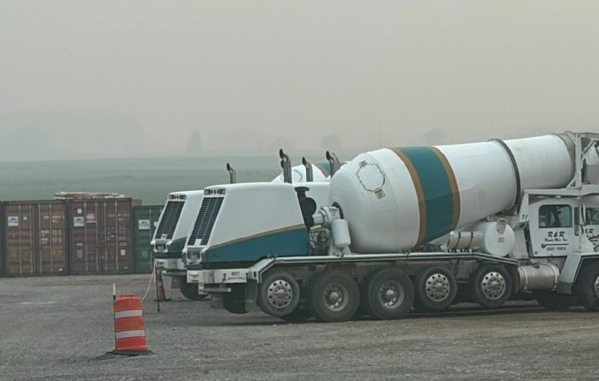 Three white cement mixer trucks with teal and gold stripes parked in a row on a gravel surface near orange traffic barrel and stacked shipping containers in foggy distance.