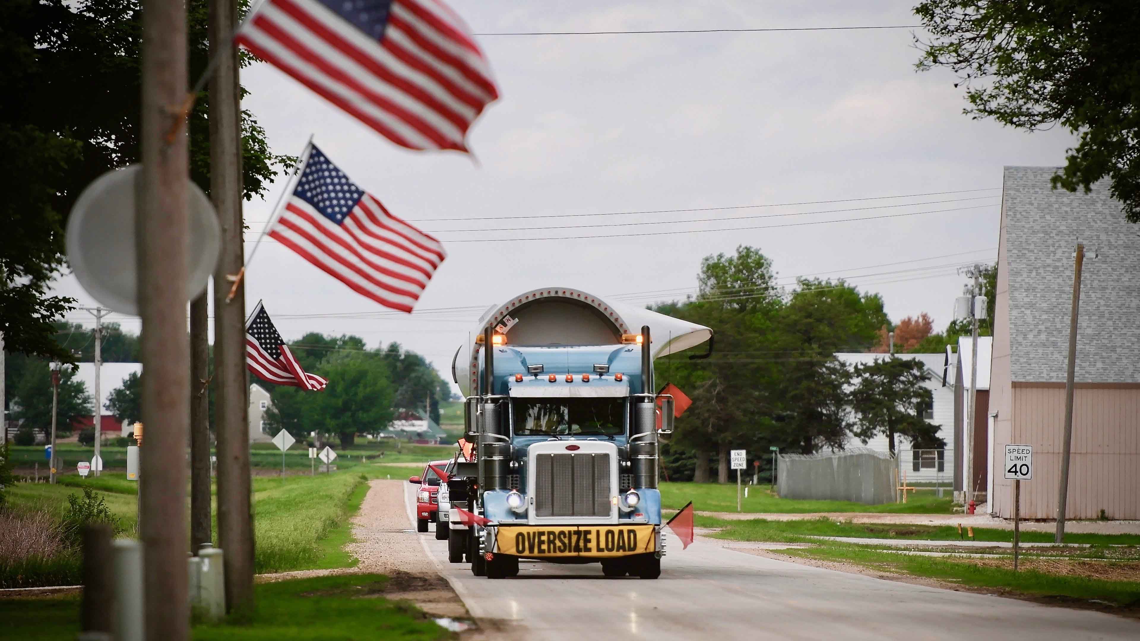A large blue semi-truck carrying a wind turbine blade marked with an 'OVERSIZE LOAD' banner drives down a rural road with American flags attached to roadside poles.