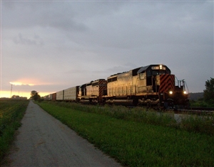 Diesel locomotive train traveling on tracks beside a paved path through grassy fields during dusk with cloudy skies and fading sunlight.
