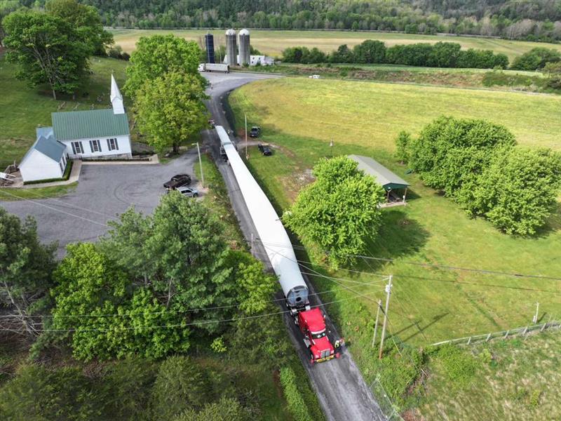 Aerial view of a long red truck transporting a large wind turbine blade on a narrow rural road passing a white church with a green roof and surrounded by green fields and trees.