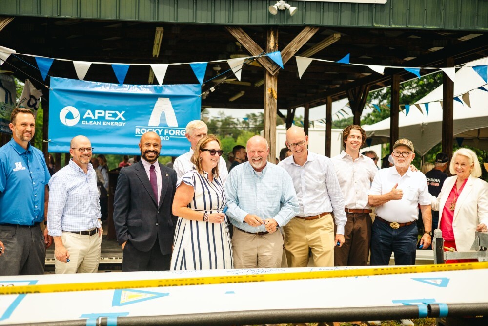 Group of men and women standing outdoors in front of a pavilion with blue and white triangular flags overhead and a banner displaying Apex Clean Energy and Rocky Forge Wind logos, smiling and posing for a photo.