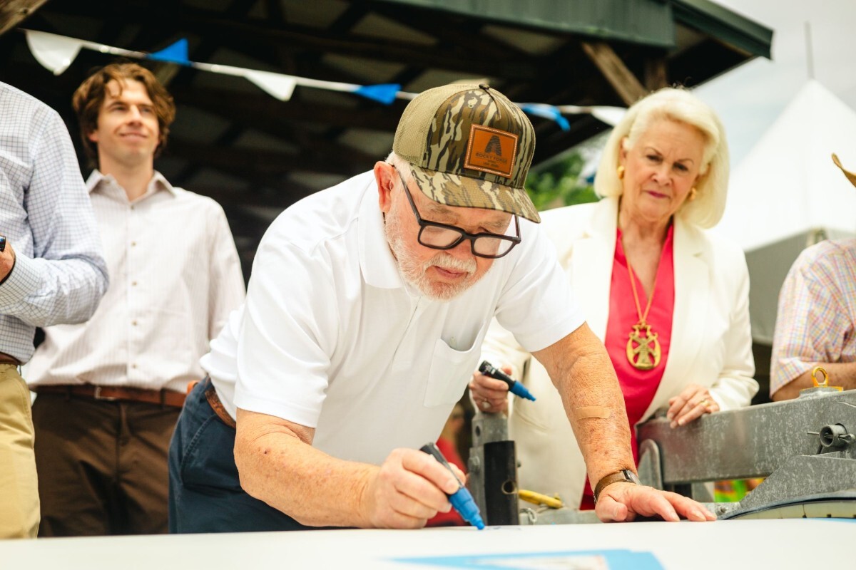 Older man with glasses and a camouflage hat writing or drawing on a white surface with a blue marker while a woman in a white blazer and red blouse watches attentively behind him, with other people and an outdoor shelter in the background.