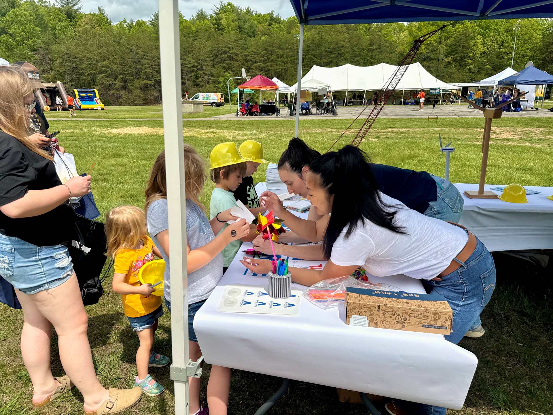 Children and adults interact at an outdoor science or engineering activity table under a tent. Two children wearing yellow construction helmets focus on a craft project with colorful pinwheels, assisted by two women leaning over the table. Additional people and event tents are visible in the grassy background under a partly cloudy sky.