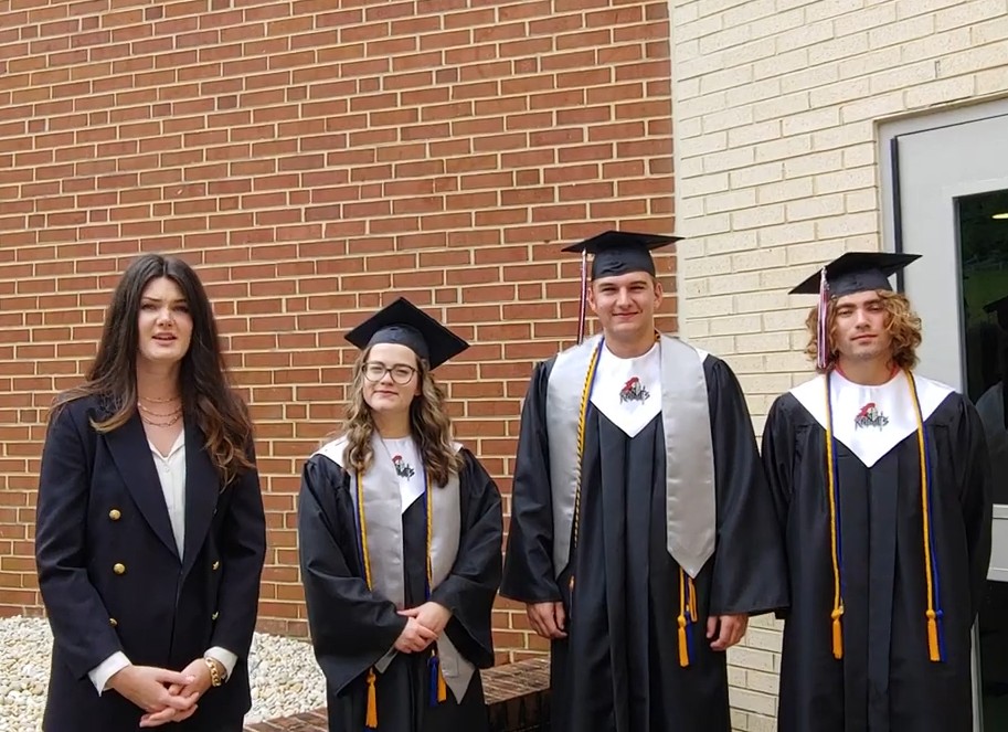 Three students in black graduation gowns and caps stand next to a woman in a black blazer in front of brick and light-colored walls.