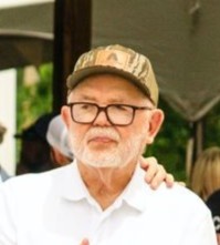 Older man wearing glasses and a beige camo-patterned baseball cap, dressed in a white shirt, with a hand resting on his shoulder.