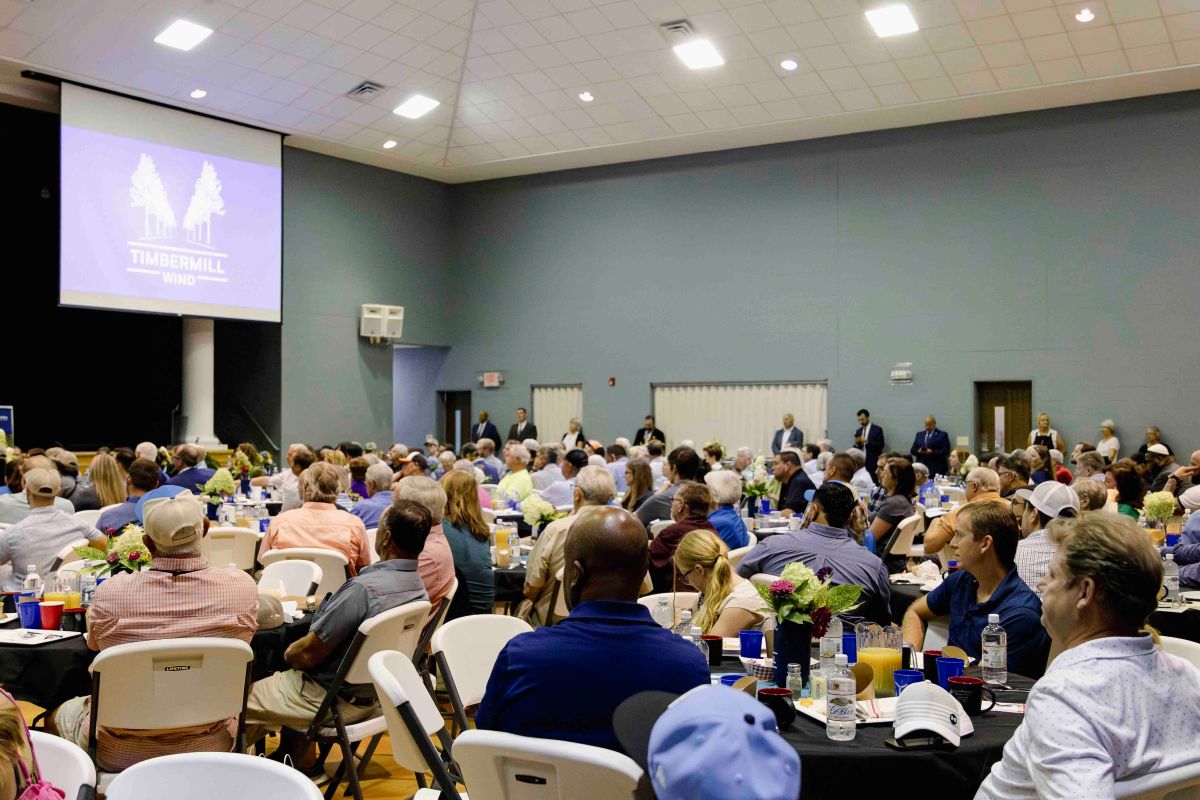 Crowd of people seated at round tables in a large hall facing a screen displaying a Timbermill Wind logo, attending an indoor event or presentation.