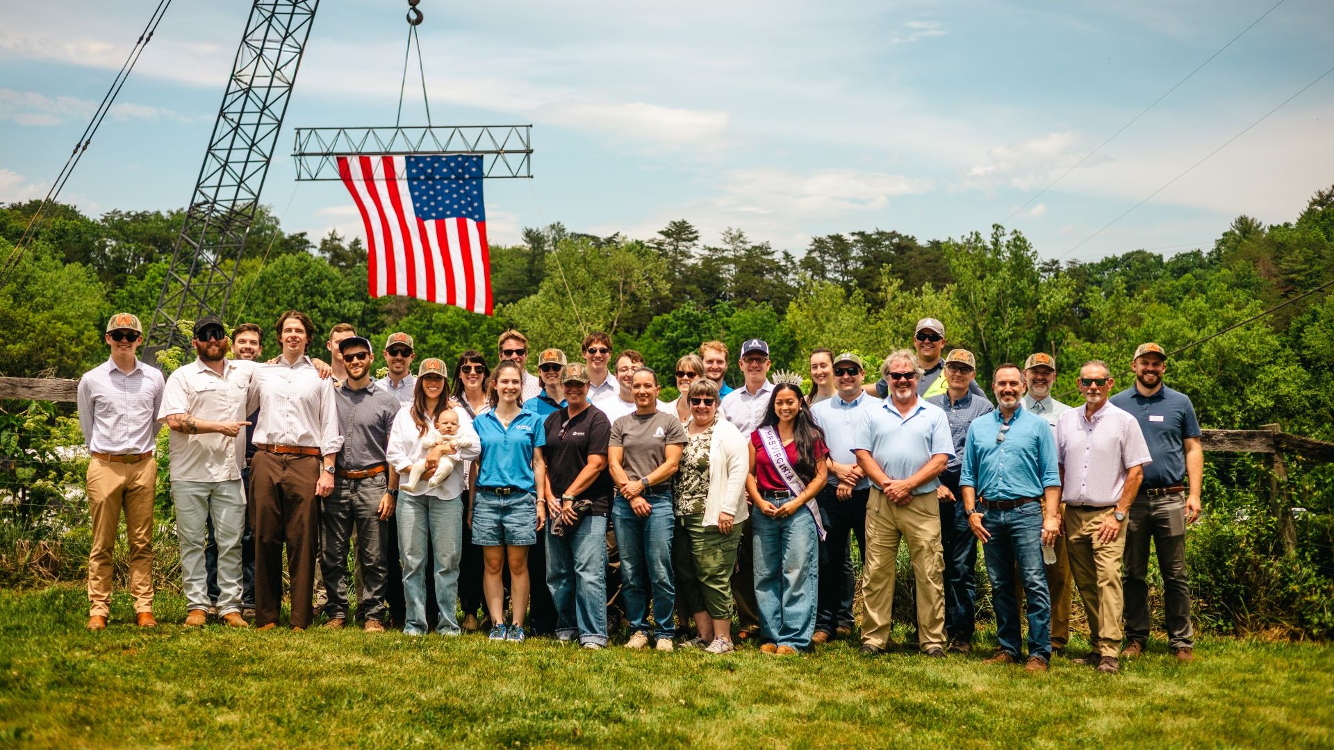 Group of about 28 people standing on grass outdoors with trees behind them, under a blue sky, posing for a photo with an American flag hanging from a crane structure above them.