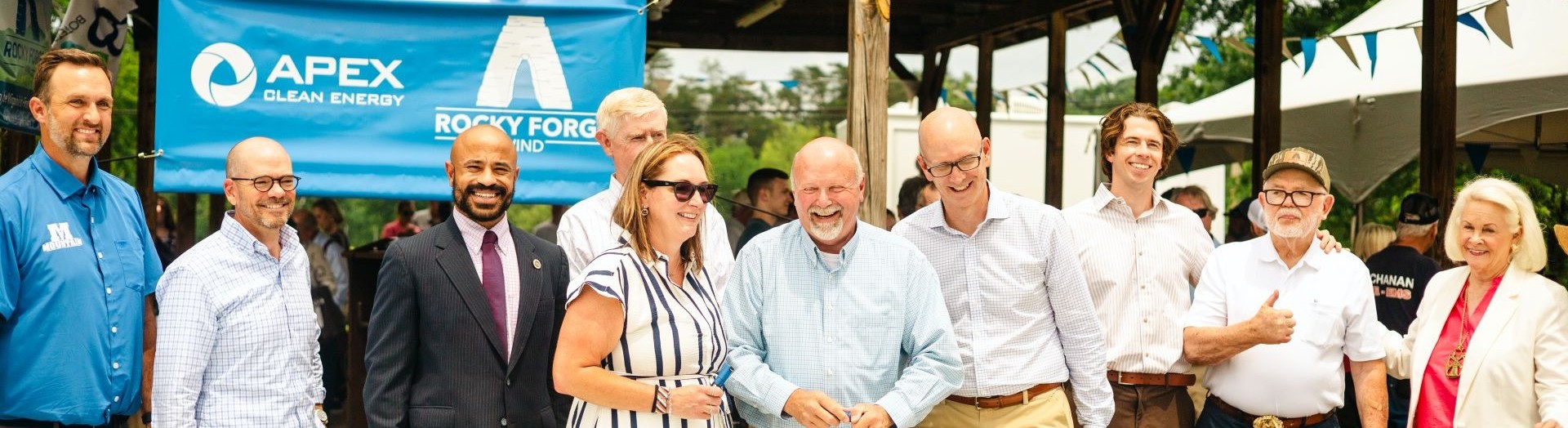 Group of nine people standing outdoors under a pavilion at an event, smiling and posing for a photo, with a blue Apex Clean Energy and Rocky Forge Wind banner in the background.