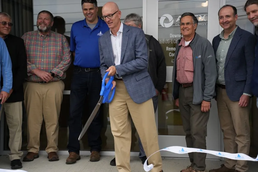 Man in a blue blazer and beige pants cutting a ceremonial ribbon with large blue scissors while a group of men stand behind him smiling outside a building with glass doors.